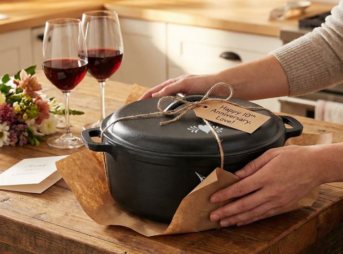 Couple gifting a personalized cast iron Dutch oven with engraved lid and Happy 10th Anniversary Love tag, set on a kitchen counter with wine glasses and flowers