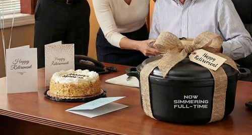 Engraved cast iron Dutch oven reading Now Simmering Full Time presented as a retirement gift beside a cake and retirement cards on an office table