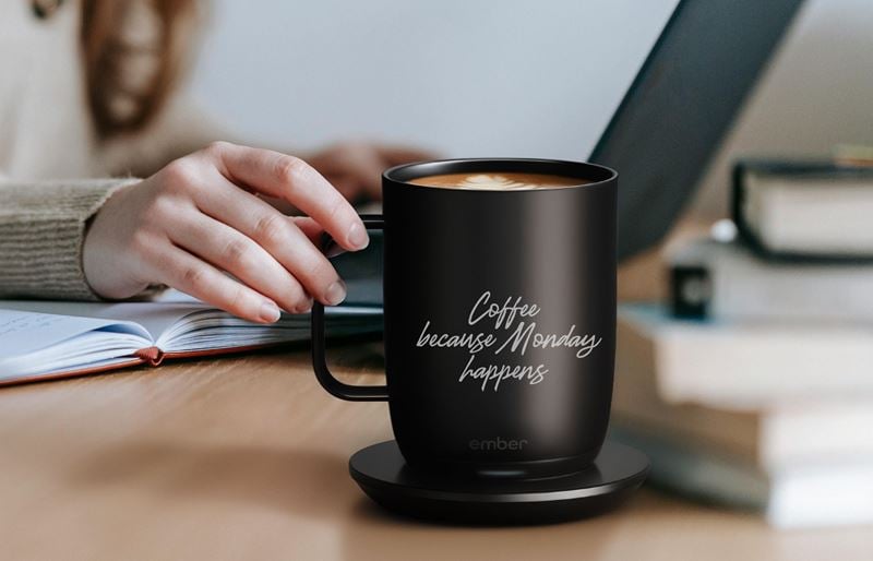 Laser engraved black Ember smart heated coffee mug on charging coaster with ‘Coffee because Monday happens’ design next to a person working at a desk