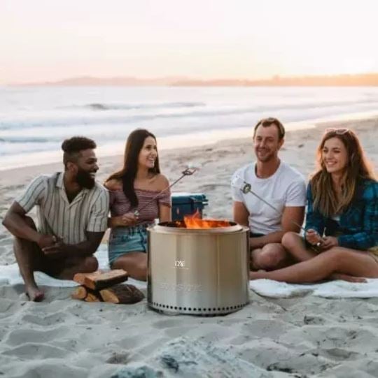 fun group of adults sitting on a beach surrounding a solo stove and roasting marshmallows